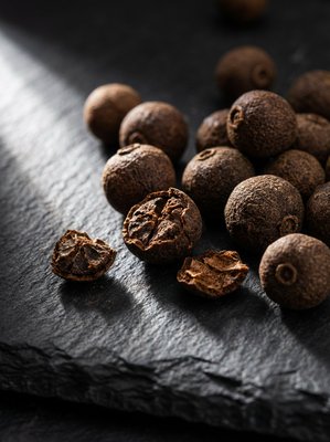 [400×400px | 72dpi | JPG] Macro food photography: Jamaican allspice berries on dark slate, dark brown spherical berries resembling peppercorns but larger, some whole and some cracked open, dramatic raking side light revealing texture and color depth, editorial macro style, no text