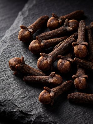 [400×400px | 72dpi | JPG] Macro food photography: whole organic cloves arranged on dark slate showing the distinctive nail-like shape with bulbous bud and long stem, deep mahogany brown color, dramatic side lighting highlighting form, ultra-sharp detail, editorial style, no text