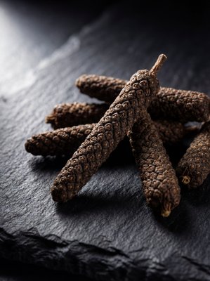 [400×400px | 72dpi | JPG] Macro food photography: long pepper (Piper longum) showing distinctive elongated catkin-like spike form, dark brown with visible seed clusters along the shaft, arranged on dark slate, dramatic lighting showing the unusual texture, editorial macro style, no text
